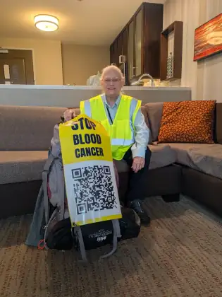 Jeana Moore in a hi-vis vest, smiling on a sofa beside her packed rucksack and tent, holding a yellow STOP BLOOD CANCER banner with a QR code on it.