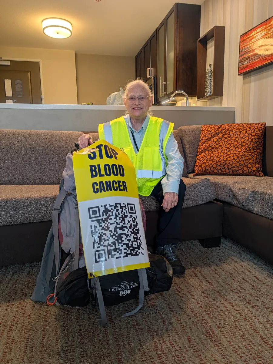 Jeana Moore in a hi-vis vest, smiling on a sofa beside her packed rucksack and tent, holding a yellow STOP BLOOD CANCER banner with a QR code on it.