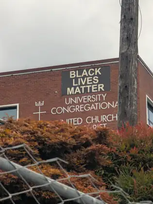 A red-brick building seen over a bent chain-link fence and red-leafed shrubs, a dark banner reading BLACK LIVES MATTER above the lettering University Congregational, beside a weathered wooden utility pole.
