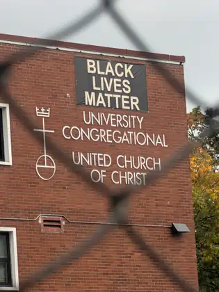Seen through a chain-link fence, a red-brick wall carries a dark banner reading BLACK LIVES MATTER above the white lettering University Congregational, United Church of Christ, and a cross emblem topped by a crown.