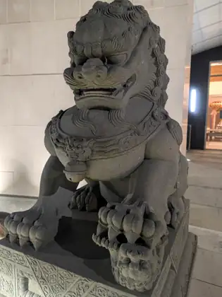A dark grey carved stone lion with a curled mane and bared teeth sits on an ornate plinth, one front paw resting on a small cub, beneath a lit covered entrance with pale stone walls.