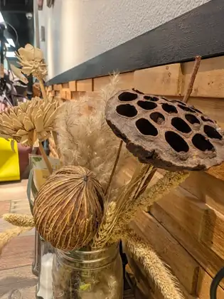 A glass jar of dried decorations, including a dark seed head pitted with deep holes, a ribbed ball-shaped pod, pale curled-wood flowers, feathery grass plumes and grain stalks, against a wall of wooden blocks.