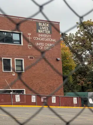 Seen through a chain-link fence, a red-brick building with a dark banner reading BLACK LIVES MATTER above the lettering University Congregational, United Church of Christ, rises over an empty parking lot whose fence carries signs reading Reserved Parking, Sunday Mornings Only.
