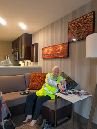 Jeana Moore, barefoot in a hi-vis vest, sits on a sofa reaching into an open zip kit of small tubes on a side table, beside a roll of tape and folded socks, under framed red artworks.