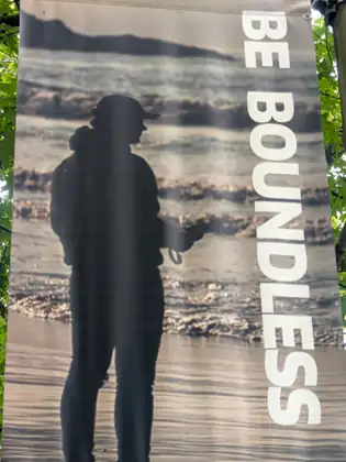 A banner hanging among green leaves shows the dark silhouette of a person in a cap standing at the edge of the surf, beside the words BE BOUNDLESS in tall white letters.