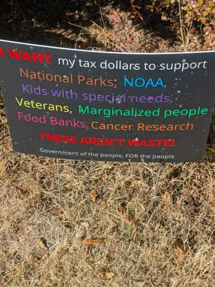 A black sign on wire stakes in dry grass reads, in multicolored letters, I WANT my tax dollars to support National Parks, NOAA, Kids with special needs, Veterans, Marginalized people, Food Banks, Cancer Research, THESE AREN’T WASTE! Government of the people, FOR the people.