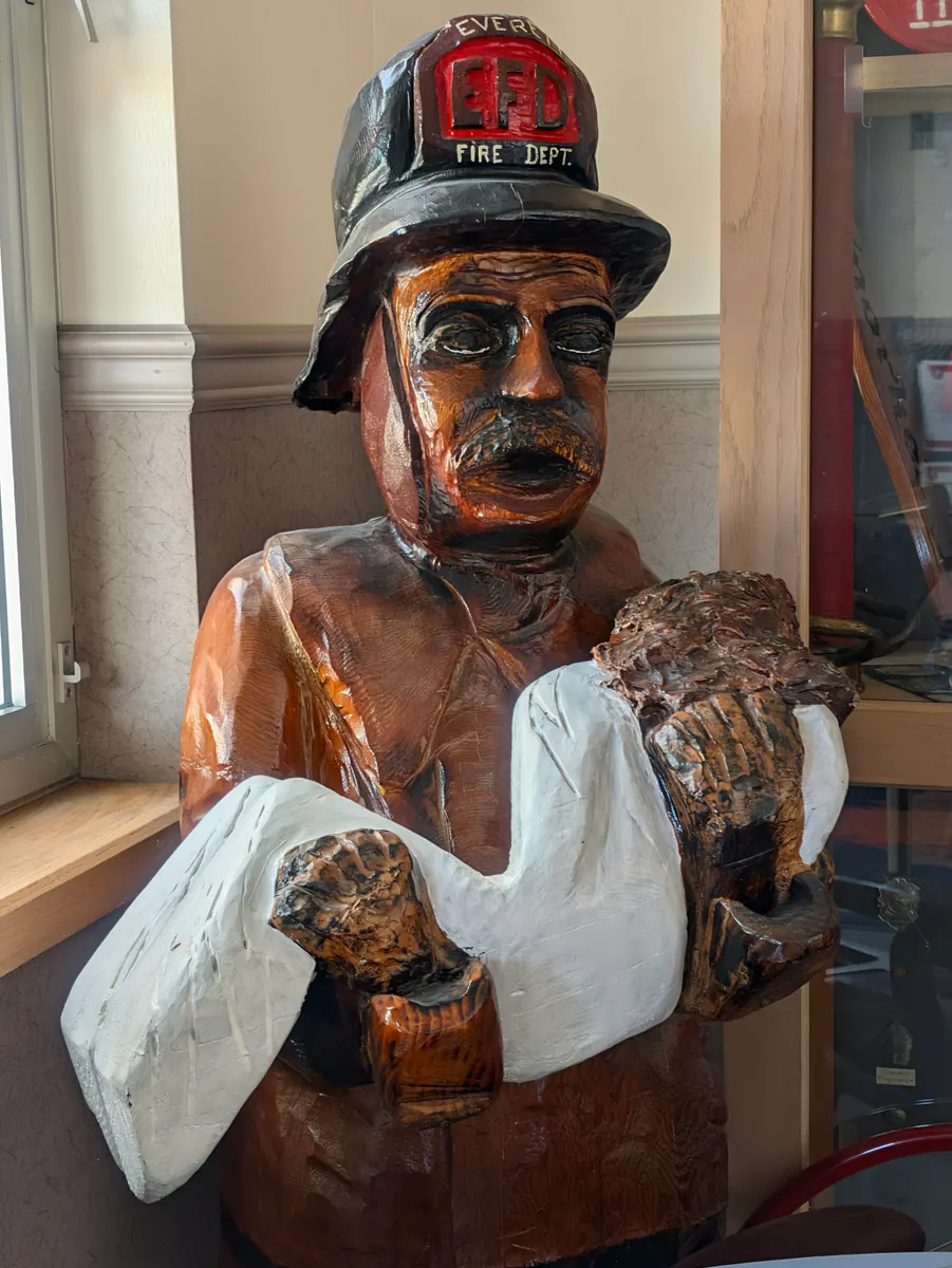 A life-size carved wooden figure of a firefighter in a red helmet, holding a bundle, standing indoors by a window.