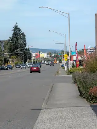 A wide arterial road on the edge of town in afternoon light, traffic in the distance and a bare sidewalk in the foreground.