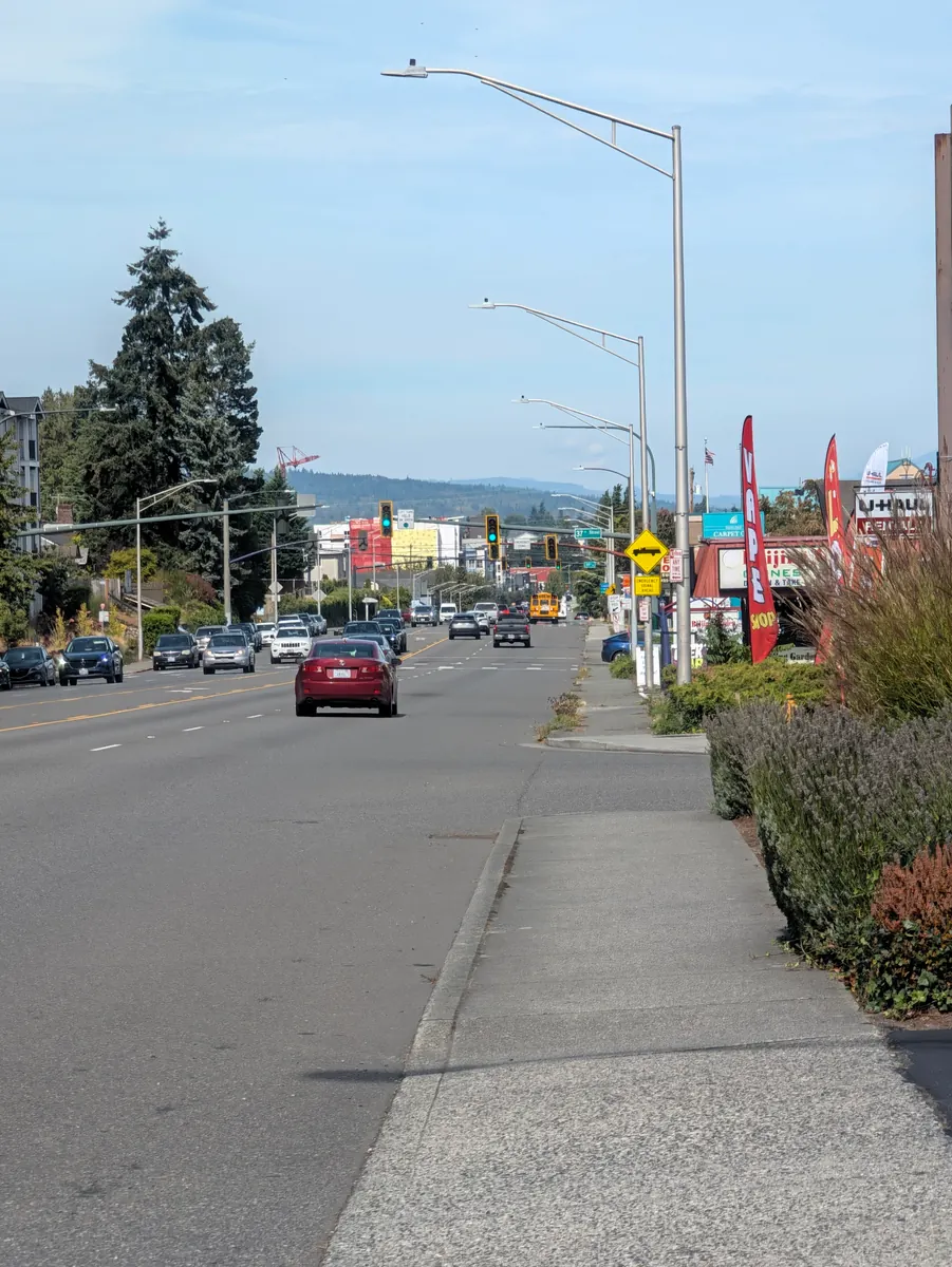A wide arterial road on the edge of town in afternoon light, traffic in the distance and a bare sidewalk in the foreground.