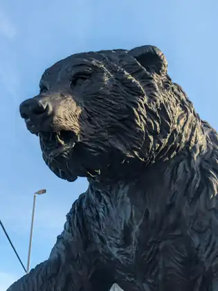 A large dark statue of a bear seen from below against a clear blue sky, its mouth half open to show its teeth, with a street lamp on a tall pole behind it.