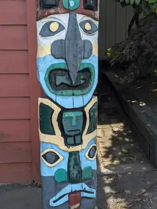 A carved wooden totem pole painted pale blue, teal, cream and grey, its faces stacked one above another, standing against red wooden siding beside a shaded brick path.