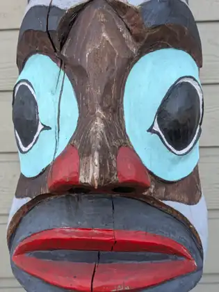 A close view of a carved wooden face on a pole, with large pale blue eyes, flared red nostrils and full red lips, the weathered wood split by long cracks.