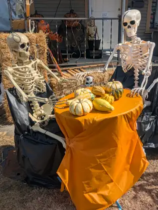 Two plastic skeletons seated at a small table draped in orange plastic and piled with pumpkins and gourds, in front of a porch decorated with straw bales and pumpkin-headed figures.
