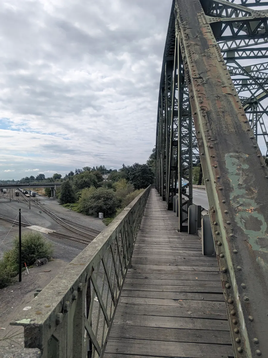 A timber walkway running along the side of a green steel truss bridge, with railway tracks and marshland below.