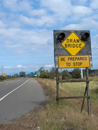 A weathered wooden board holding a yellow sign reading Draw Bridge and Be Prepared to Stop between two signal lights, beside a road that runs on towards a steel bridge and green highway signs.