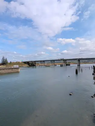 A long concrete highway bridge crosses the far end of a wide river, trucks travelling along it, with a timber-sided pier on the left and weathered pilings and rocks along the right edge.