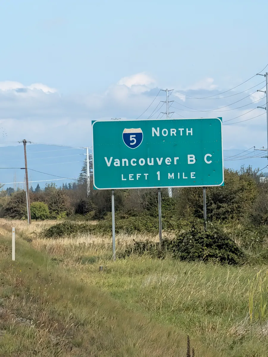 A green highway sign reading Interstate 5 North, Vancouver B C, Left 1 Mile, standing in rough grass under a wide sky.