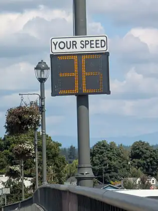 A speed display reading YOUR SPEED shows 36 in amber lights on a tall grey pole above a bridge railing, beside an old-style street lamp and hanging flower baskets, with rooftops, trees and distant mountains beyond.
