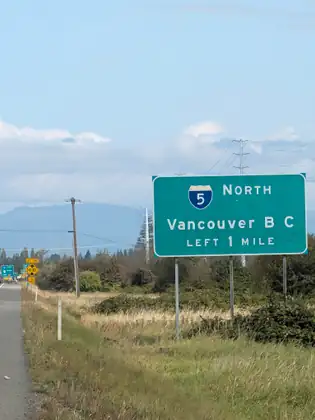 A green highway sign reading Interstate 5 North, Vancouver B C, Left 1 Mile, stands in rough grass beside a road shoulder that runs ahead toward yellow signs reading Left Lane, 14'-10", 1/2 Mile, with power lines and distant hills under a blue sky.