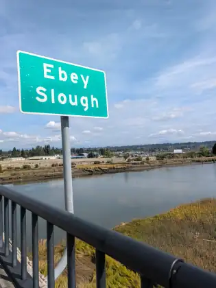 A road sign reading Ebey Slough beside a bridge railing, with tidal water and low buildings beyond.