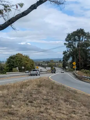 A highway curves down past a dry grassy bank towards two green steel truss bridges, with a white pickup and other cars on the road and yellow signs reading Overheight Detector Ahead and 14'-0" at the roadside.
