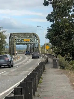 Cars cross a road bridge through a green steel truss arch marked 14 FT 0 IN, beside a yellow sign reading Overheight Stop When Flashing, with a narrow footpath running along the timber barrier on the right.