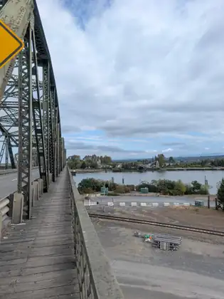 A weathered plank walkway runs along the outside of a green riveted truss bridge, looking down over the railing at a railway line, an empty gravel lot and a river with wooded banks beyond.
