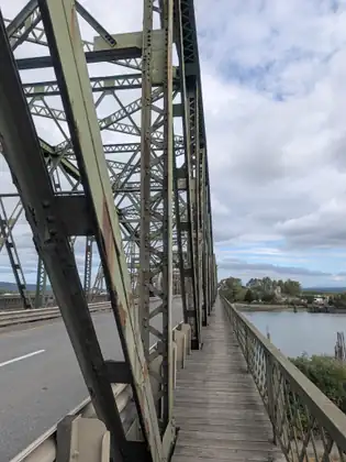 Looking up at the rusting green girders of a steel truss bridge, with an empty roadway on the left and a long timber walkway running straight ahead beside the river on the right.
