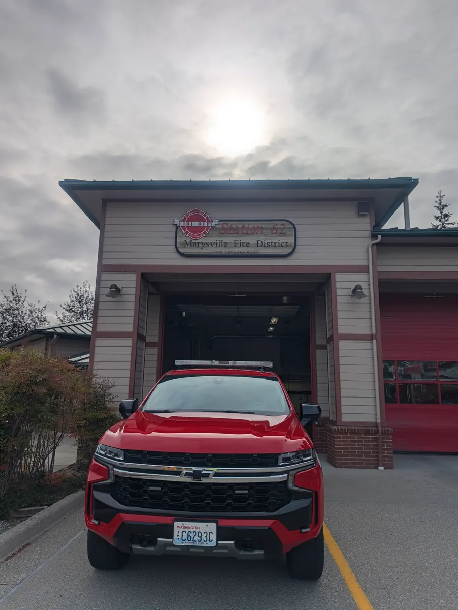 The front of a fire station under a bright overcast sky, with a red command truck parked in the open bay door.