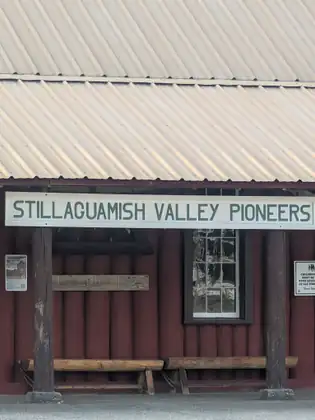 The red log front of a building under a pale ribbed metal roof, with a long sign reading Stillaguamish Valley Pioneers, a carved board reading Pioneer Hall 1923 and two log benches.