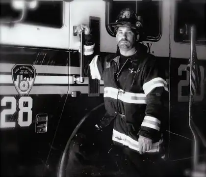 An archival black-and-white photograph of a firefighter in a helmet and turnout coat with reflective stripes, one hand on a grab bar, standing beside a fire engine marked 28 with a Fire Department City of New York emblem on its side.
