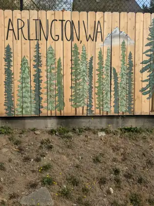 A wooden fence painted with a row of fir trees and a mountain, lettered Arlington, WA.
