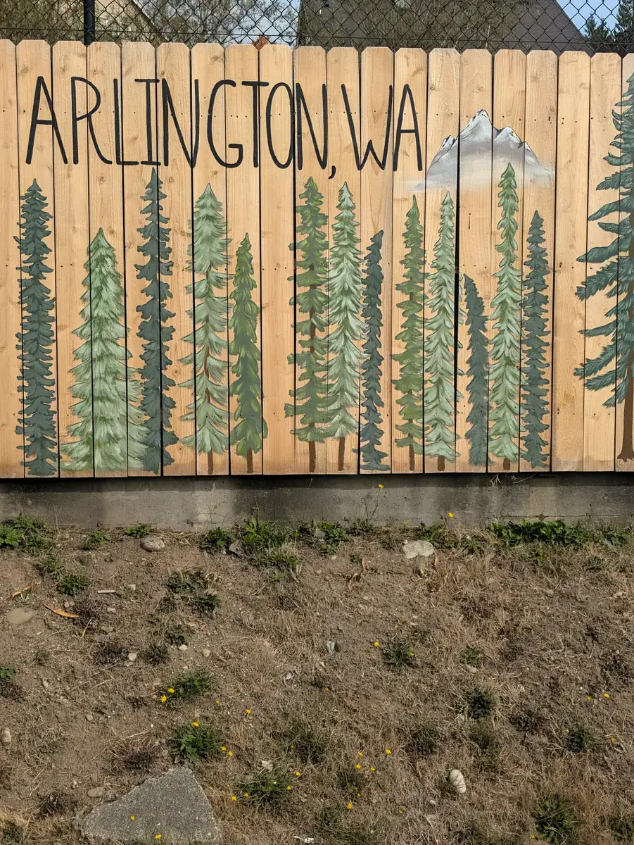 A wooden fence painted with a row of fir trees and a mountain, lettered Arlington, WA.