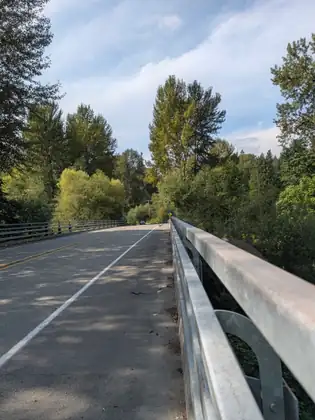 A two-lane road crossing a bridge, a galvanised steel railing running along the near side, with tall trees ahead under a blue sky and one car far off.