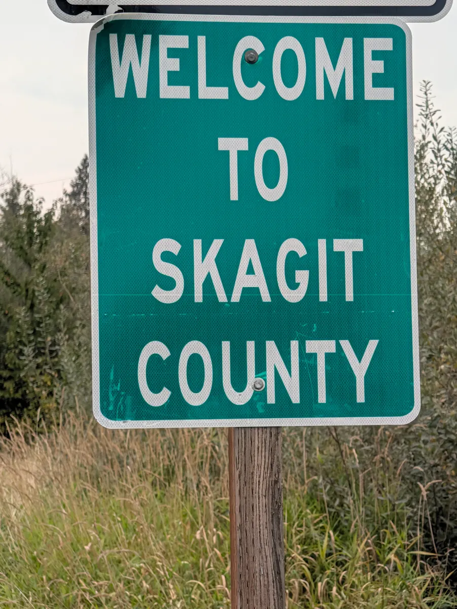 A green road sign reading Welcome to Skagit County, against trees and a pale sky.