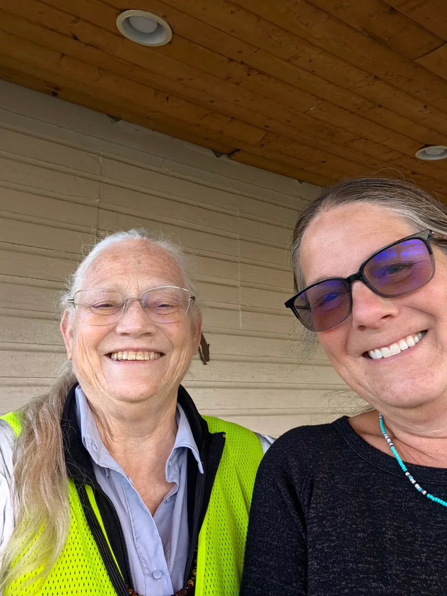 Jeana Moore in her hi-vis vest, smiling shoulder to shoulder with Becky under the eaves of a porch.