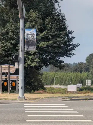 A painted crosswalk across an empty road, a metal pole with a pedestrian signal and a banner painted with a locomotive, a weathered timber frame on the left, and a hand-lettered sign reading G-Sale in the verge beyond.