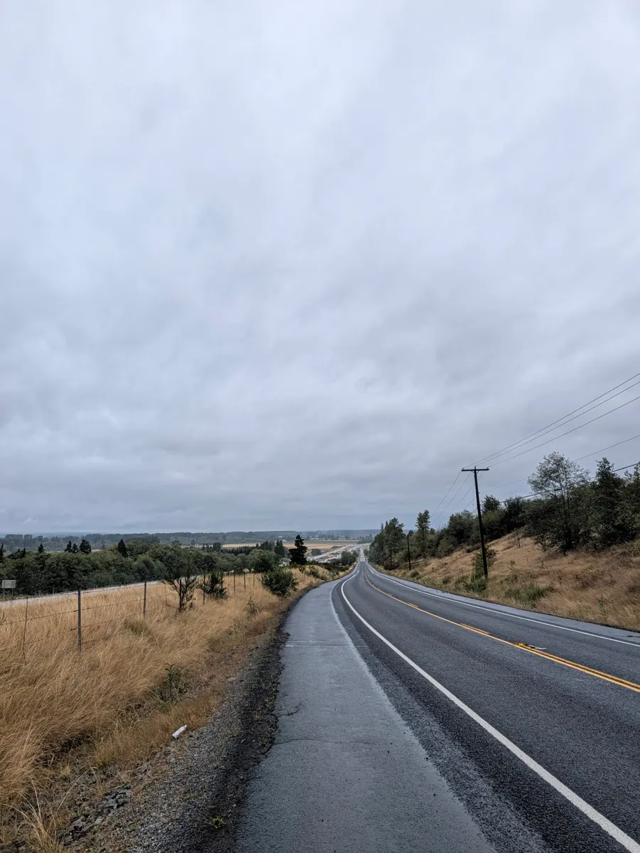 A wet two-lane road running away between fields under a low grey sky, the tarmac shining with rain.