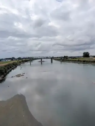 A wide, still river seen from a bridge, with an old steel truss bridge crossing it in the distance under heavy cloud.