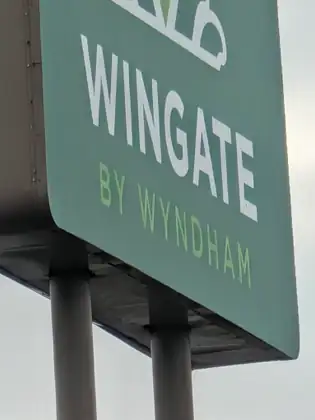 Looking up from below at the corner of a tall green sign on two gray poles, its white and light green lettering reading WINGATE BY WYNDHAM beneath part of a white logo, against a flat gray sky.