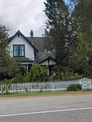 A white Victorian house with steep gables and a porch, set back behind a white picket fence beside the road.