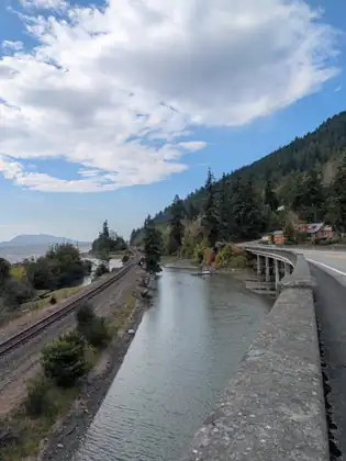 A railway line and a still channel running side by side below the road, with a bridge crossing and a forested hill beyond.