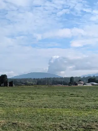 A long mountain ridge with a tall column of cloud standing above it, over mown green fields and farm buildings.