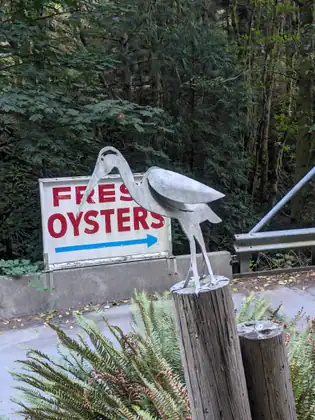 A hand-painted sign reading Fresh Oysters with an arrow, beside a life-size metal heron standing on a post.