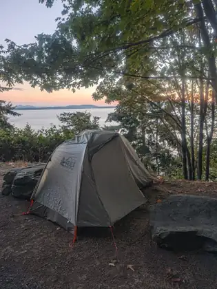 A small grey dome tent pitched on bare ground between two boulders, the bay and the last of the sunset showing through the trees behind it.