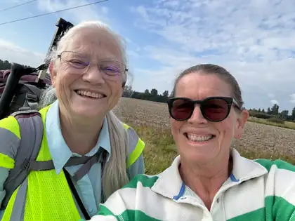 Jeana Moore in her hi-vis vest and pack straps, smiling cheek to cheek with a woman in sunglasses and a green and white striped shirt, a bare ploughed field behind them.