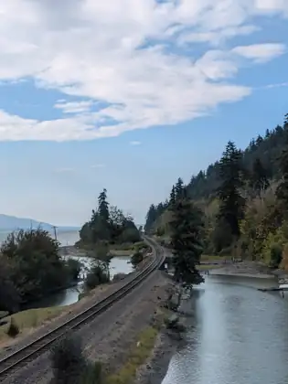 A railroad track curving along a wooded shoreline between a still channel and a small lagoon, with old pilings on the mudflat, a small wooden float and the open bay beyond.
