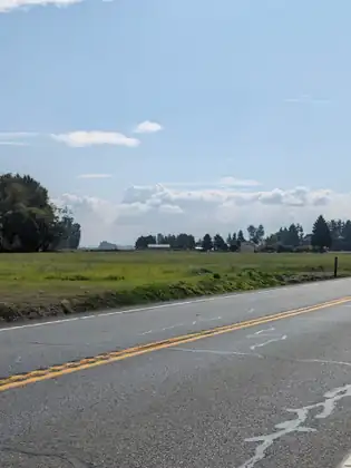 An empty two-lane road with a yellow centre line, running past open fields under a pale sky.