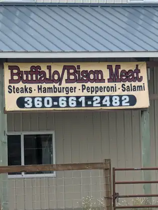 A hand-lettered sign on a metal barn reading Buffalo and Bison Meat, steaks, hamburger, pepperoni, salami, with a phone number.