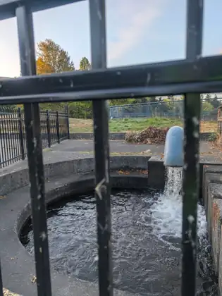 Water pouring from a pale blue pipe into a round concrete pool, seen through black iron railings beside Whatcom Creek.